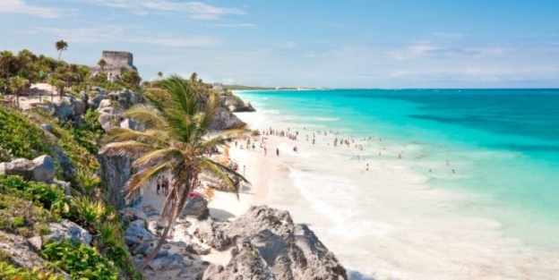 A beach with palm trees and blue water with Tulum in the background AI-generated content may be incorrect.